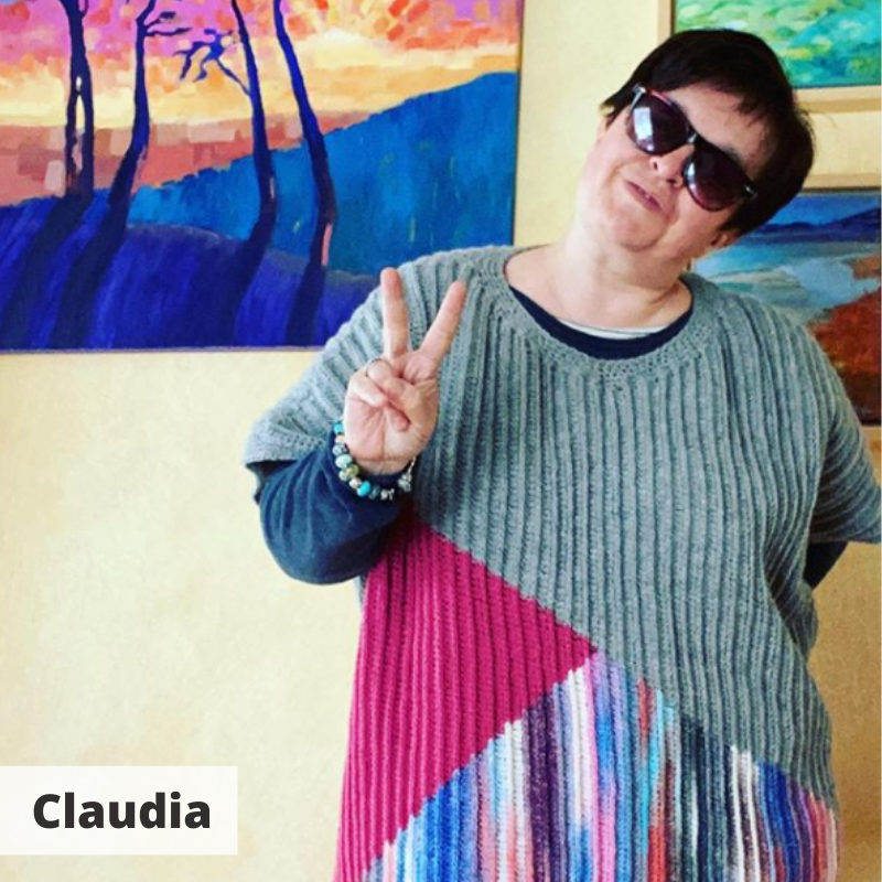 Claudia in her house wearing sunglasses and her crocheted acre sweater, head tilted to one side and doing a peace sign.